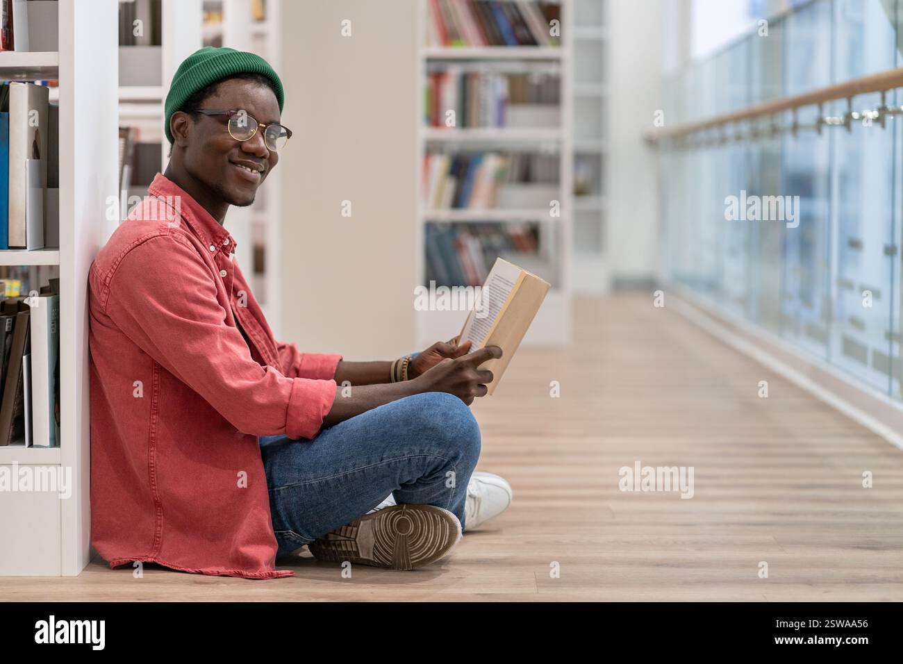 Sorridente Nero studente uomo lettura libro in biblioteca universitaria , guardando la macchina fotografica. Hobby di lettura. Foto Stock