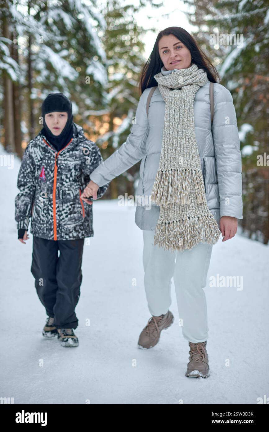 Fotografia di una foresta innevata con una donna e un ragazzo che camminano mano nella mano. La donna, posizionata in primo piano, ha lunghi capelli scuri Foto Stock