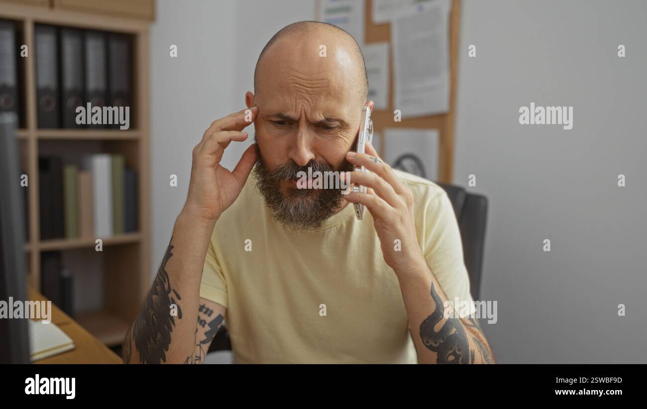 Uomo ispanico seduto in un ufficio al chiuso, sembra serio mentre parla su uno smartphone con una mano sulla testa, circondato da scaffali e documentari Foto Stock