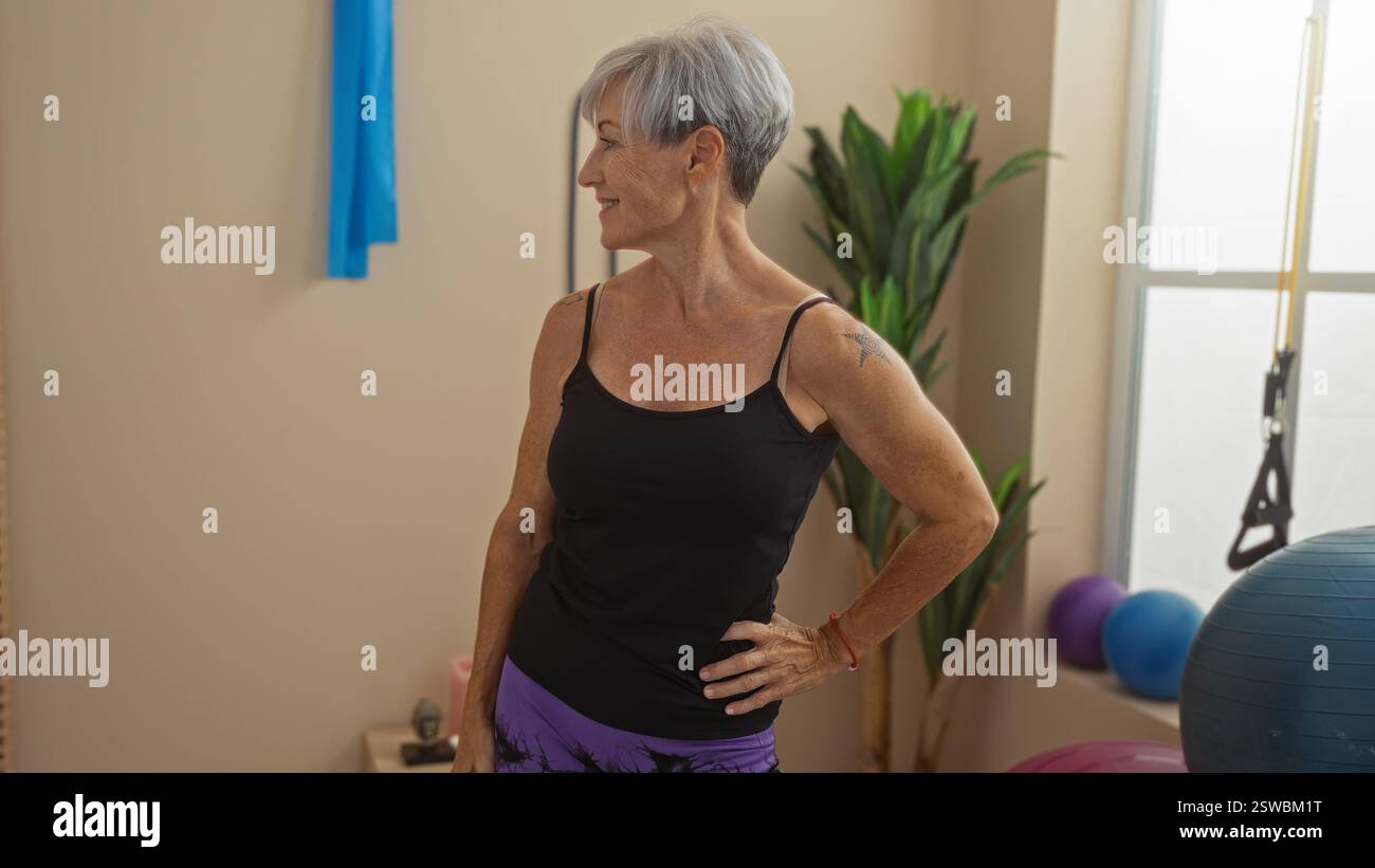 Donna di mezza età con i capelli grigi corti in palestra con attrezzature per il fitness, con canotta nera e leggings viola, sorridente e look Foto Stock