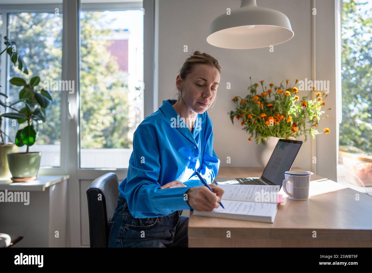 Una donna concentrata e confortevole lavora a casa scrivendo appunti nell'agenda giornaliera, seduto a un tavolo con un laptop. Foto Stock