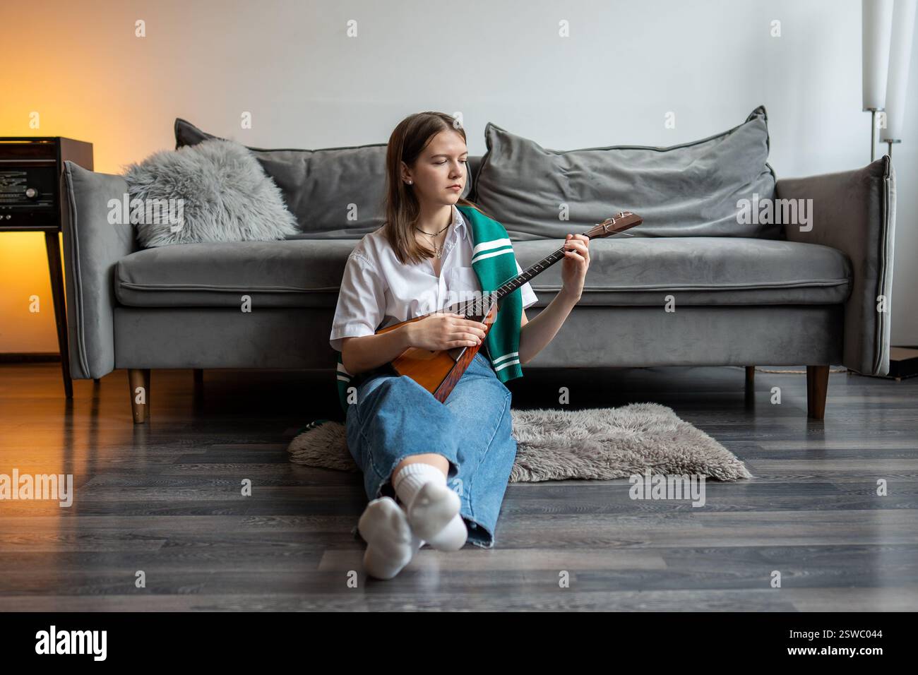 Calm Woman gioca a balalaika sul pavimento del soggiorno vicino al divano, pratica i movimenti delle dita, abilità di levigatura Foto Stock