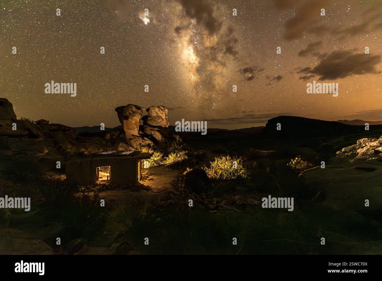 Cielo notturno della via Lattea su capanna di Adobe illuminata e formazioni rocciose nel paesaggio del deserto Foto Stock