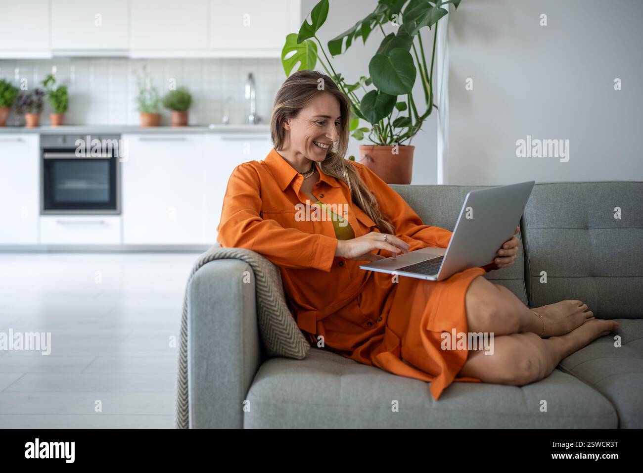 Una donna di mezza età che usa un notebook sorridente e si rilassa sul divano trascorrendo il tempo libero in una casa accogliente. Foto Stock