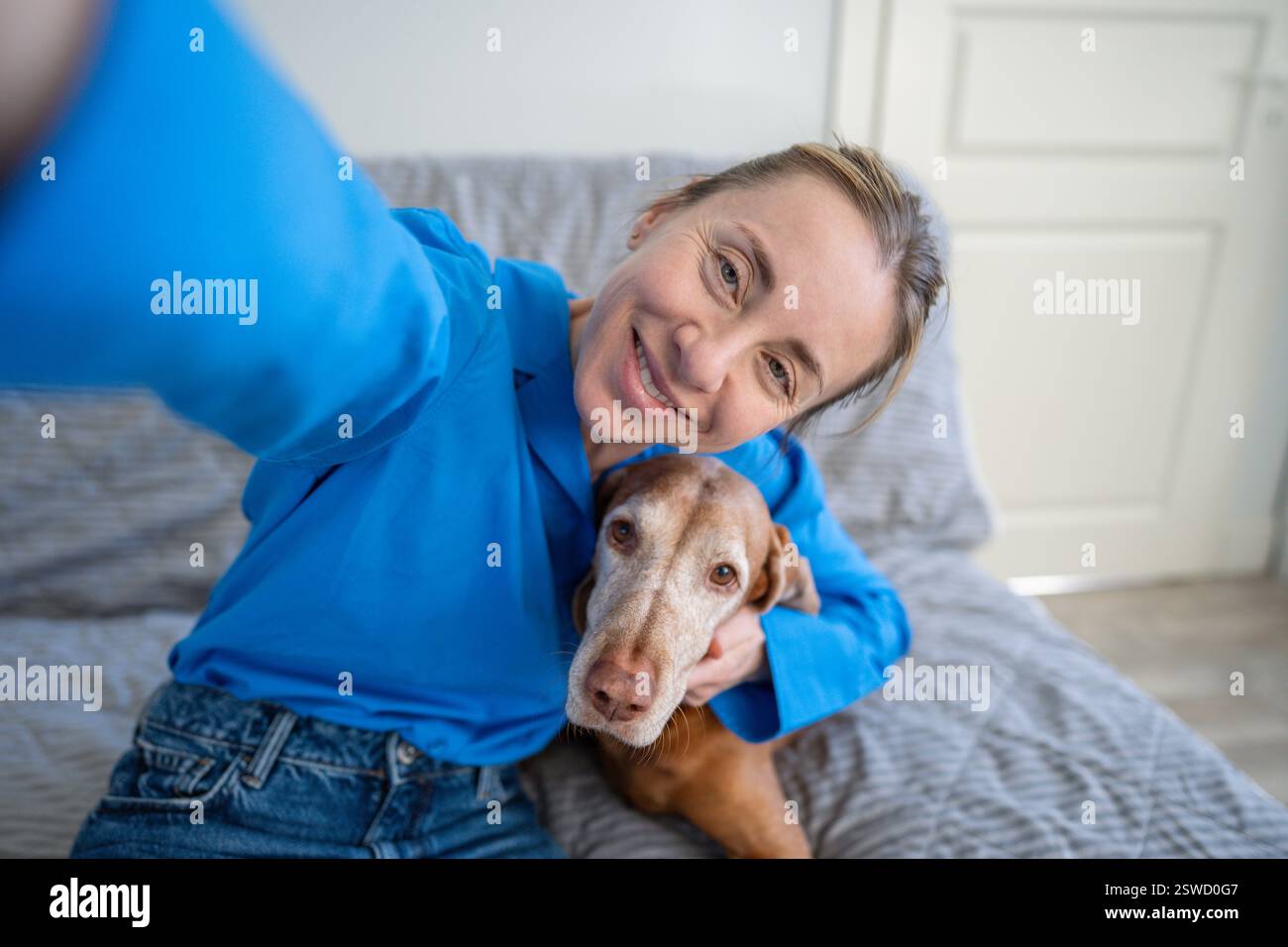 Selfie di una donna sorridente che abbraccia il cane Vizsla sul divano di casa. Affetto proprietario, salute animale domestico, amore Foto Stock