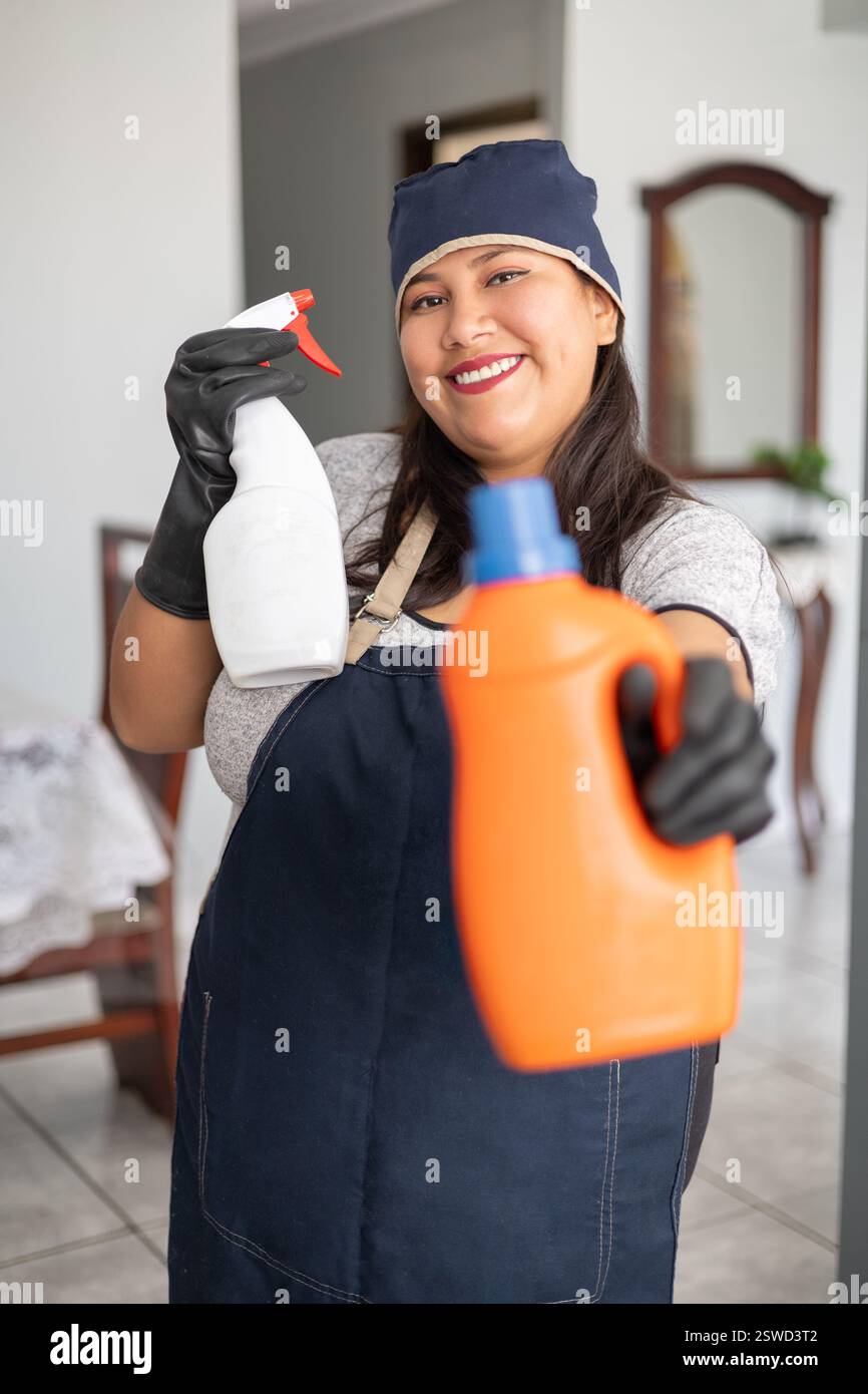 Una governante sorridente si posa con una bottiglia arancione per la pulizia in una mano e una bianca nell'altra. Indossa un grembiule blu e guanti neri, pronti a farlo Foto Stock