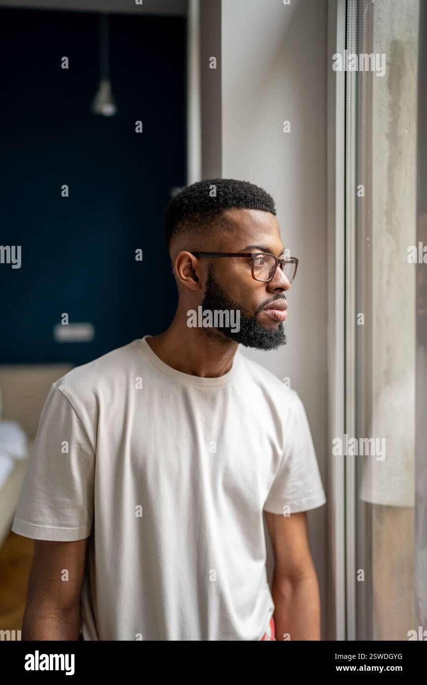 Rovesciato giovane afro-americano uomo in piedi a casa guardando fuori finestra e meditando Foto Stock