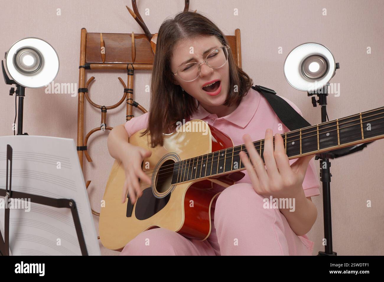 La giovane donna con gli occhiali da vista suona la chitarra acustica a sei corde e canta la canzone con tanta emozione Foto Stock