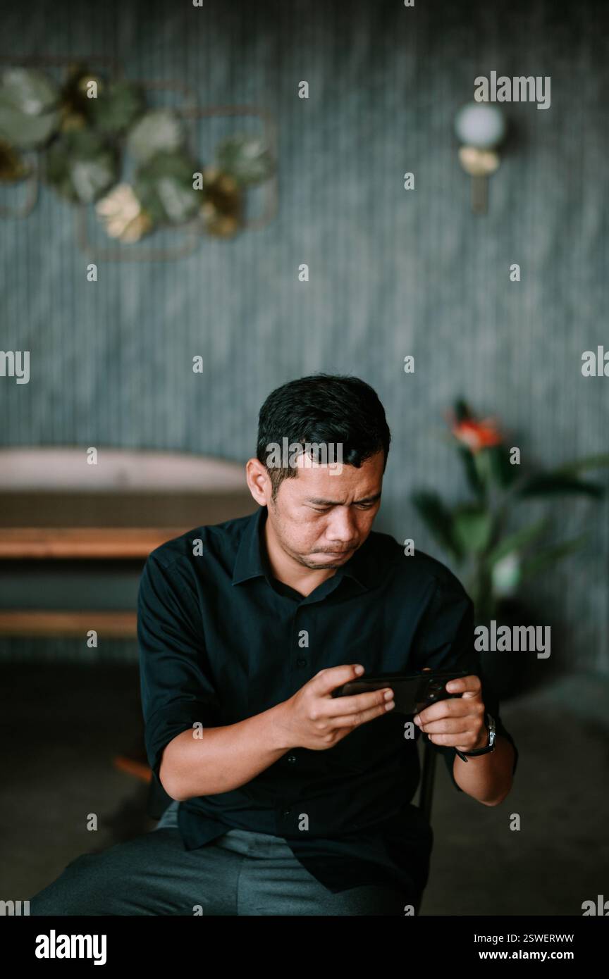 Un uomo dai capelli scuri corti, concentrato e attento, è avvolto nello schermo del suo smartphone in un ambiente interno. Indossa una camicia scura e una padella grigia Foto Stock