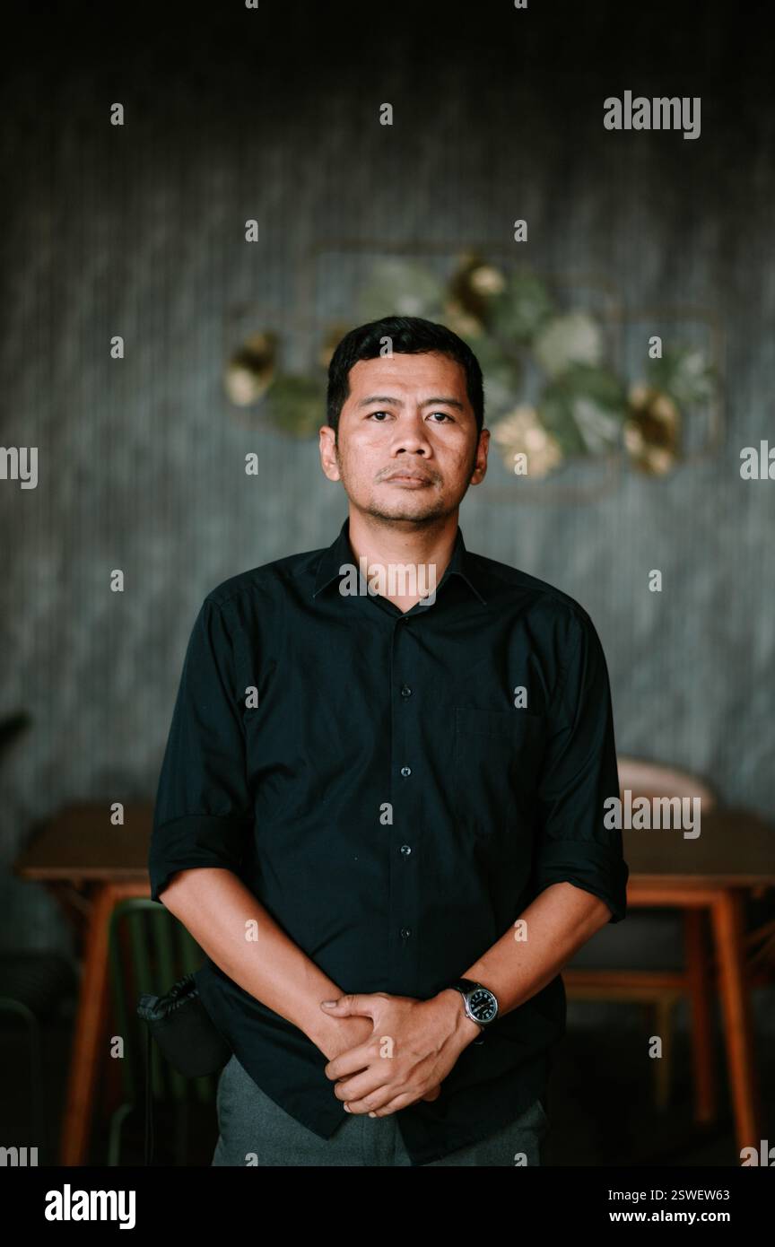 Un ritratto di un uomo indonesiano con una camicia scura, che posa al chiuso. Trasuda un comportamento serio e premuroso. Lo sfondo offre un contrasto morbido. H Foto Stock