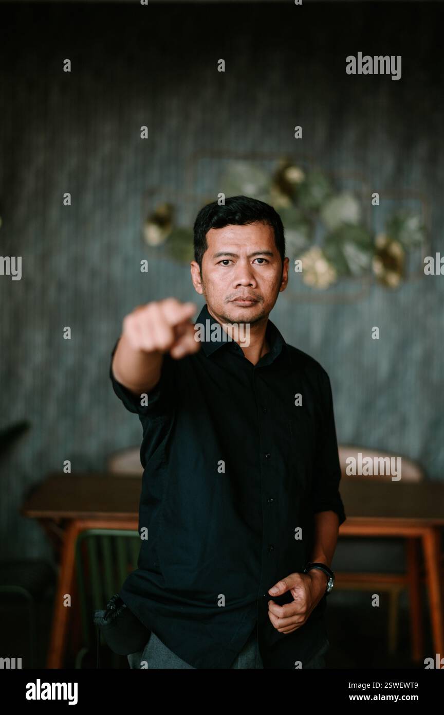 Un uomo con una camicia scura punta direttamente allo spettatore con un'espressione seria. Il gesto trasmette fiducia e senso dello scopo. Lo sfondo Foto Stock