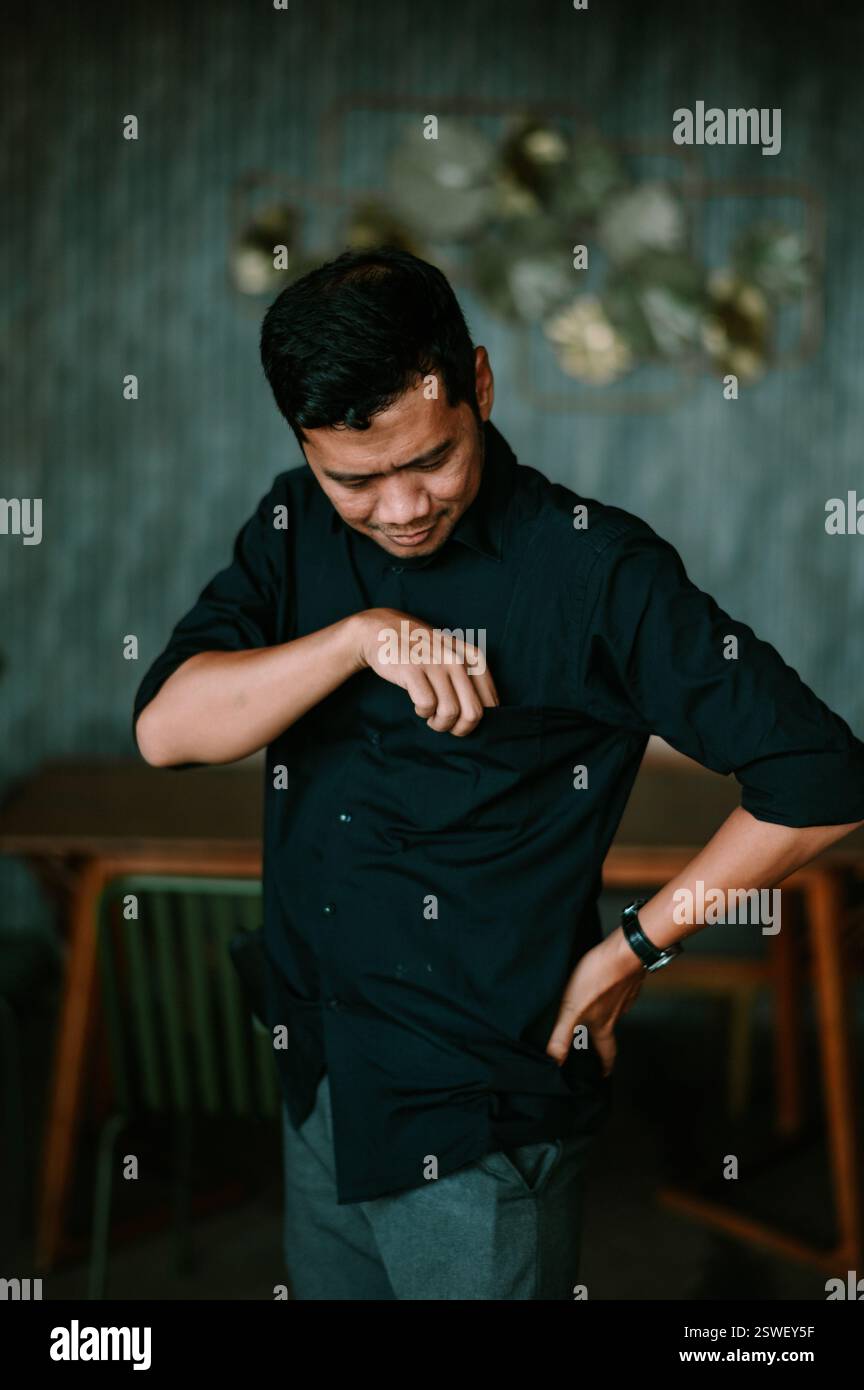 Un uomo con una camicia scura si vede controllare la tasca con uno sguardo leggermente verso il basso, apparendo premuroso. Lo sfondo è un interno silenzioso, suggerendo Foto Stock