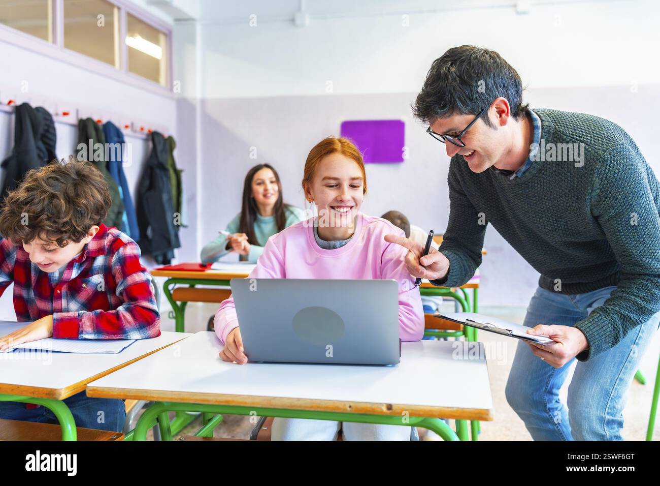 Insegnante sorridente che assiste gli studenti con un notebook in una vivace classe, promuovendo un ambiente di apprendimento di supporto per tutti Foto Stock