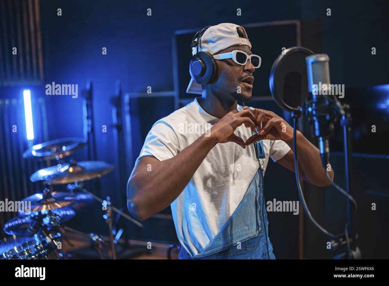 Giovane ed elegante cantante maschile che crea la forma del cuore con le mani mentre registra una nuova canzone di successo in uno studio di musica professionale Foto Stock