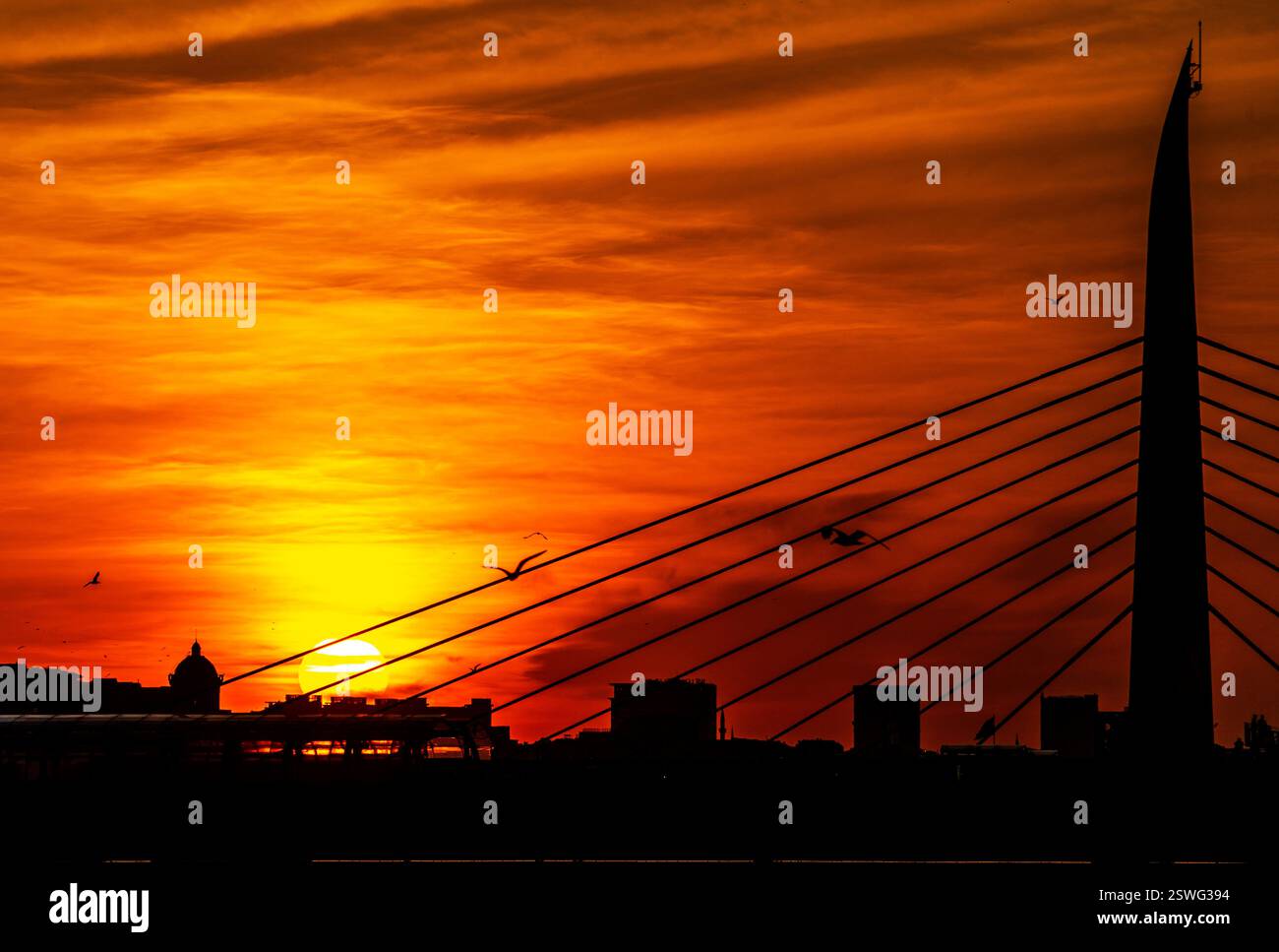 Uno splendido tramonto sullo skyline della città, caratterizzato da edifici sagomati e da un prominente ponte con cavi. Il cielo è pieno di arancione e y vibranti Foto Stock