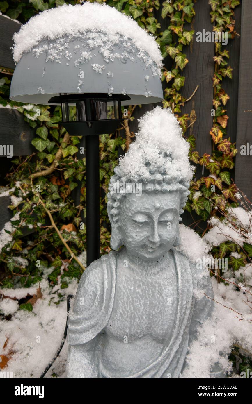 Coperte di neve, una statua di Buddha serena e una lampada in un tranquillo giardino, con edera lussureggiante che crea un'atmosfera tranquilla. Foto Stock