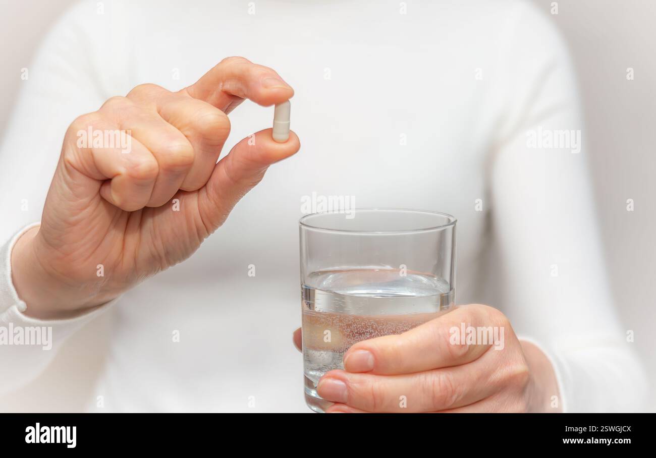 Una donna tiene una capsula in una mano e un bicchiere d'acqua nell'altra, pronta a prendere i suoi farmaci in una stanza ben illuminata. Foto Stock