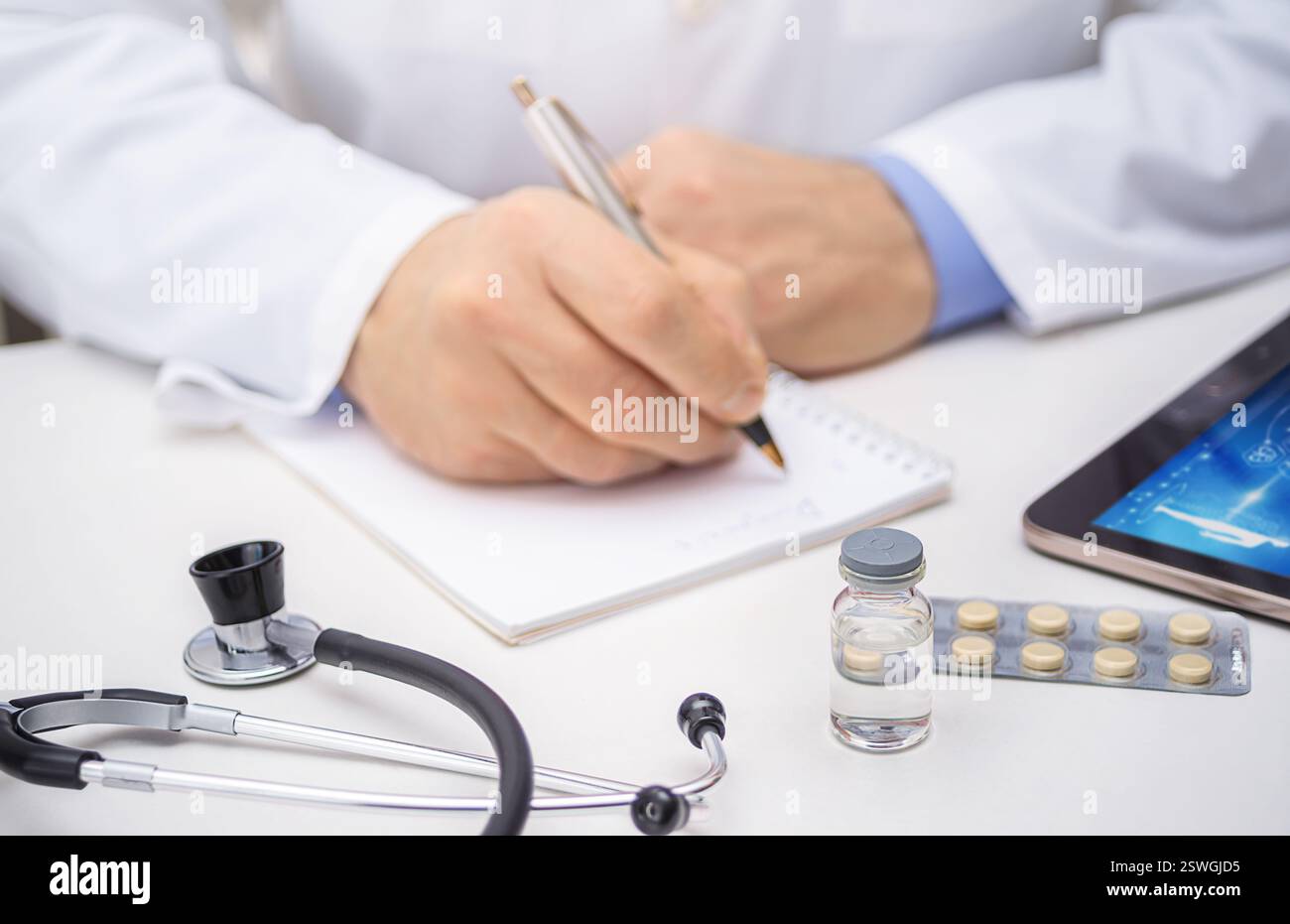 Un operatore sanitario scrive note in una cartella paziente mentre uno stetoscopio, un farmaco e una compressa si trovano nelle vicinanze, indicando una consultazione continua. Foto Stock