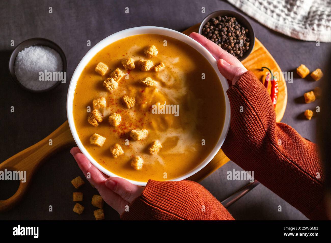 Donna che tiene una ciotola di zuppa di zucca con le zanne. Tavolo servito con zuppa panna, peperoncino e tovagliolo. Zuppa di verdure piatta Foto Stock