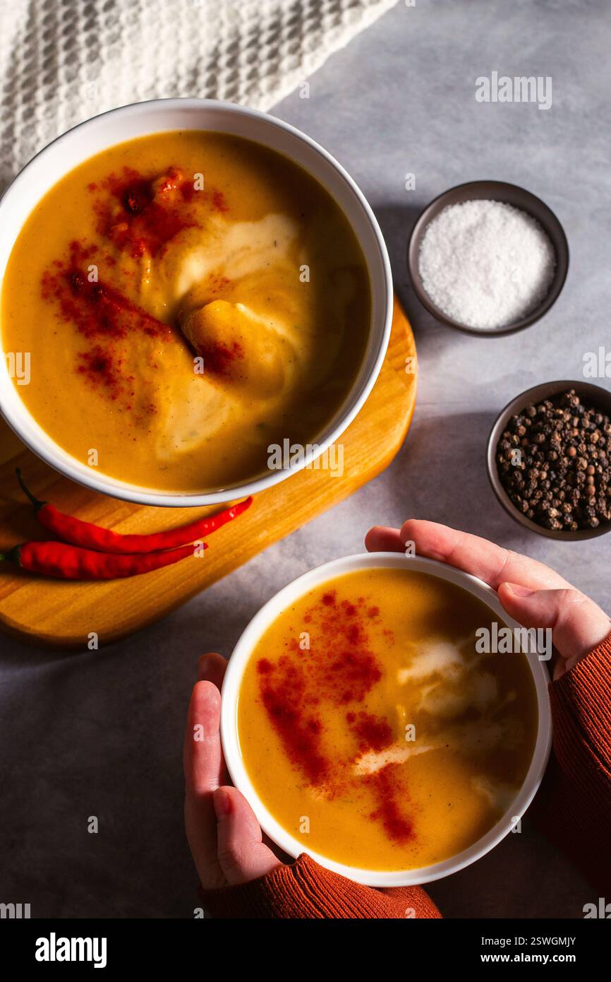 Donna che tiene una ciotola di zuppa di zucca con le zanne. Tavolo servito con zuppa panna, peperoncino e tovagliolo. Zuppa di verdure piatta Foto Stock