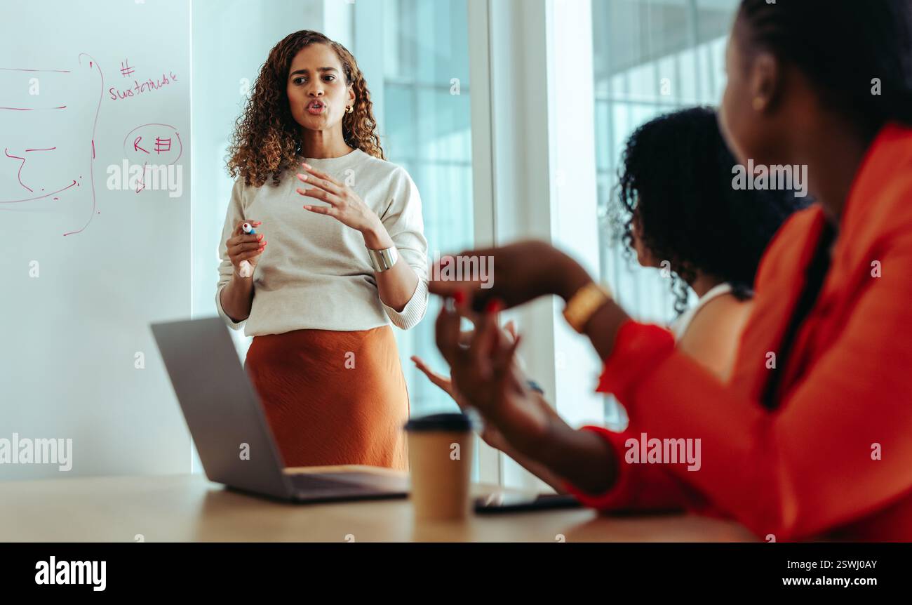Una donna d'affari sicura che presenta una strategia di marketing ai colleghi in un ambiente moderno. Si trova accanto a una lavagna con il non strategico Foto Stock