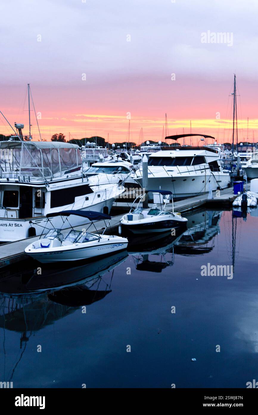 Un gruppo di barche è parcheggiato in un porticciolo, con il sole che tramonta sullo sfondo. Le barche sono di varie dimensioni e tipi, tra cui alcune di piccole dimensioni Foto Stock
