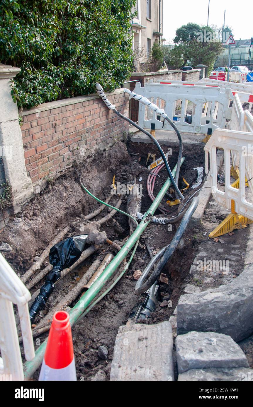 E' stata scavata una trincea per strada, probabilmente per lavori di utilità. Vari tubi e cavi, alcuni avvolti in rivestimenti protettivi, sono esposti. Foto Stock