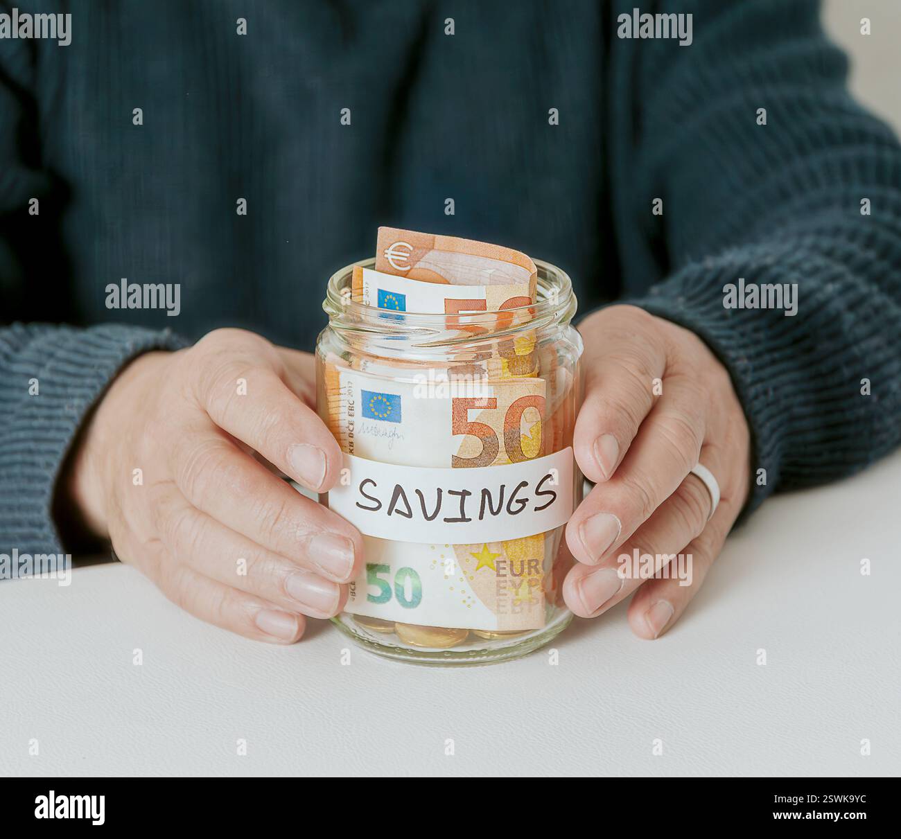 Vaso in vetro con etichetta e denaro all'interno tenuto da entrambe le mani su un tavolo bianco. Foto Stock