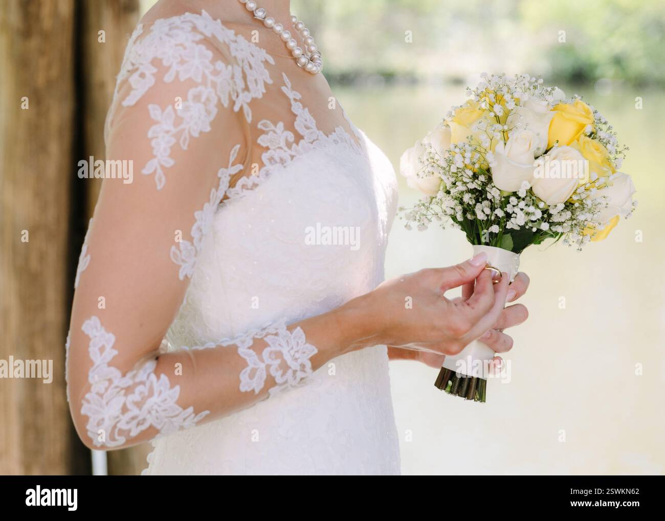 Una sposa mostra il suo bellissimo bouquet di rose bianche e gialle vicino a un tranquillo corso d'acqua, irradiando gioia e bellezza. Foto Stock