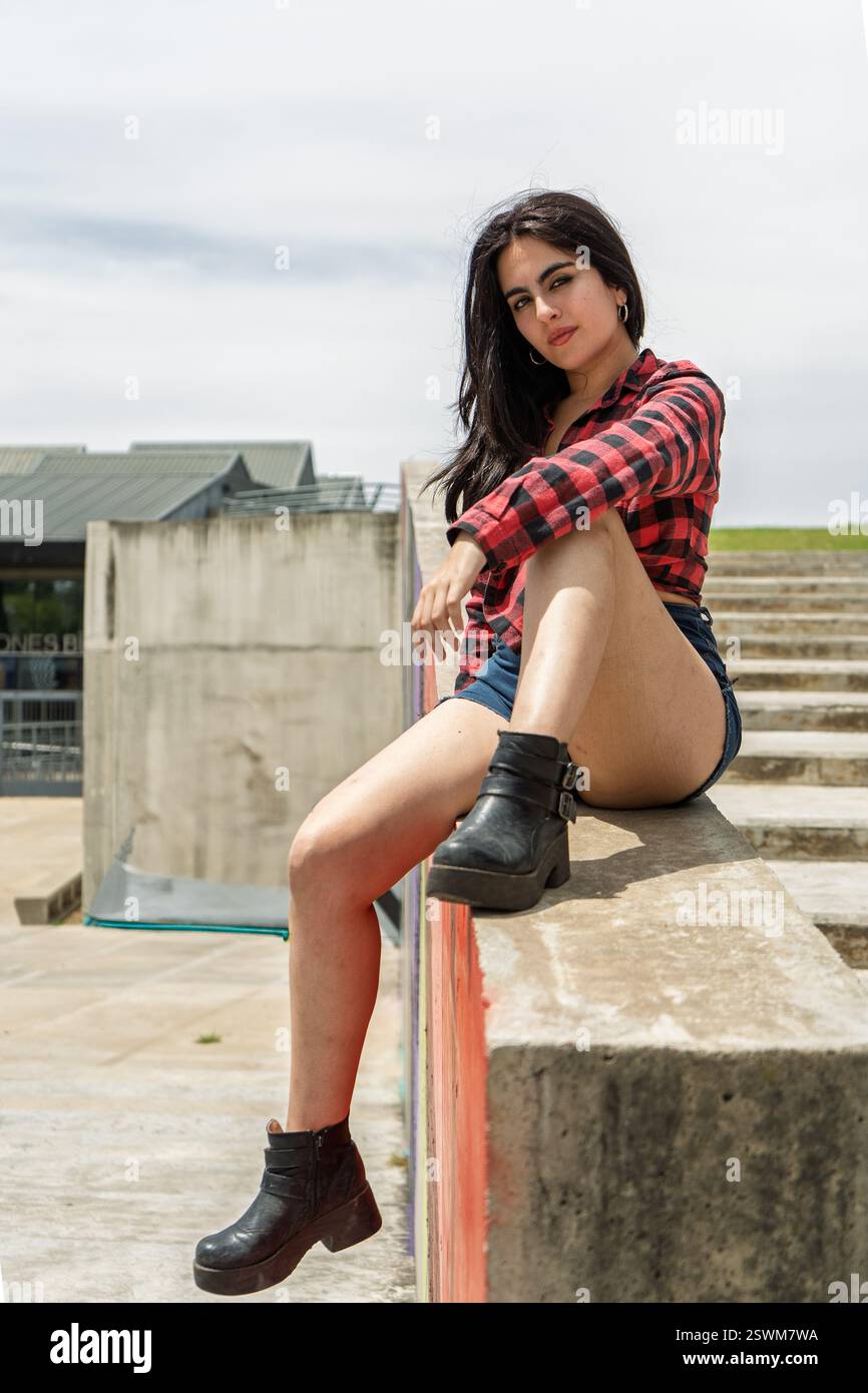 Giovane donna elegante che posa in uno skatepark, indossa una camicia a scacchi, pantaloncini in denim e stivali neri Foto Stock