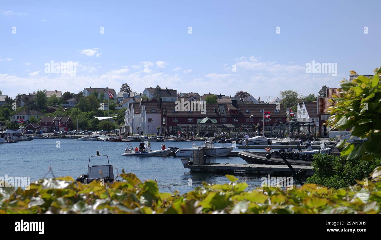 Porto di Lillesand in Norvegia decorato con bandiere per il giorno della Costituzione, con barche, case di legno e gente del posto che si gode il sole primaverile. Foto Stock