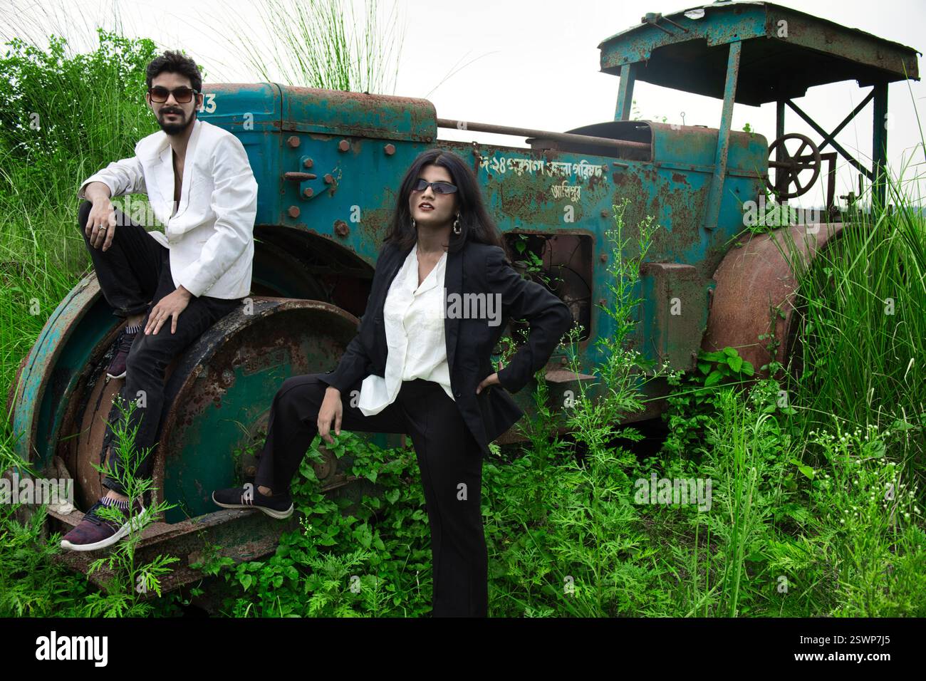 Modelli in posa in un campo aperto di fronte a un vecchio rullo. Il campo ha l'erba verde e sono vestiti in abiti bianchi e neri. Foto Stock