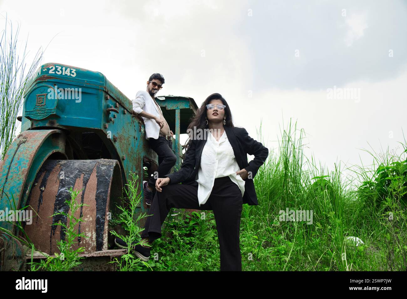 Modelli in posa in un campo aperto di fronte a un vecchio rullo. Il campo ha l'erba verde e sono vestiti in abiti bianchi e neri. Foto Stock