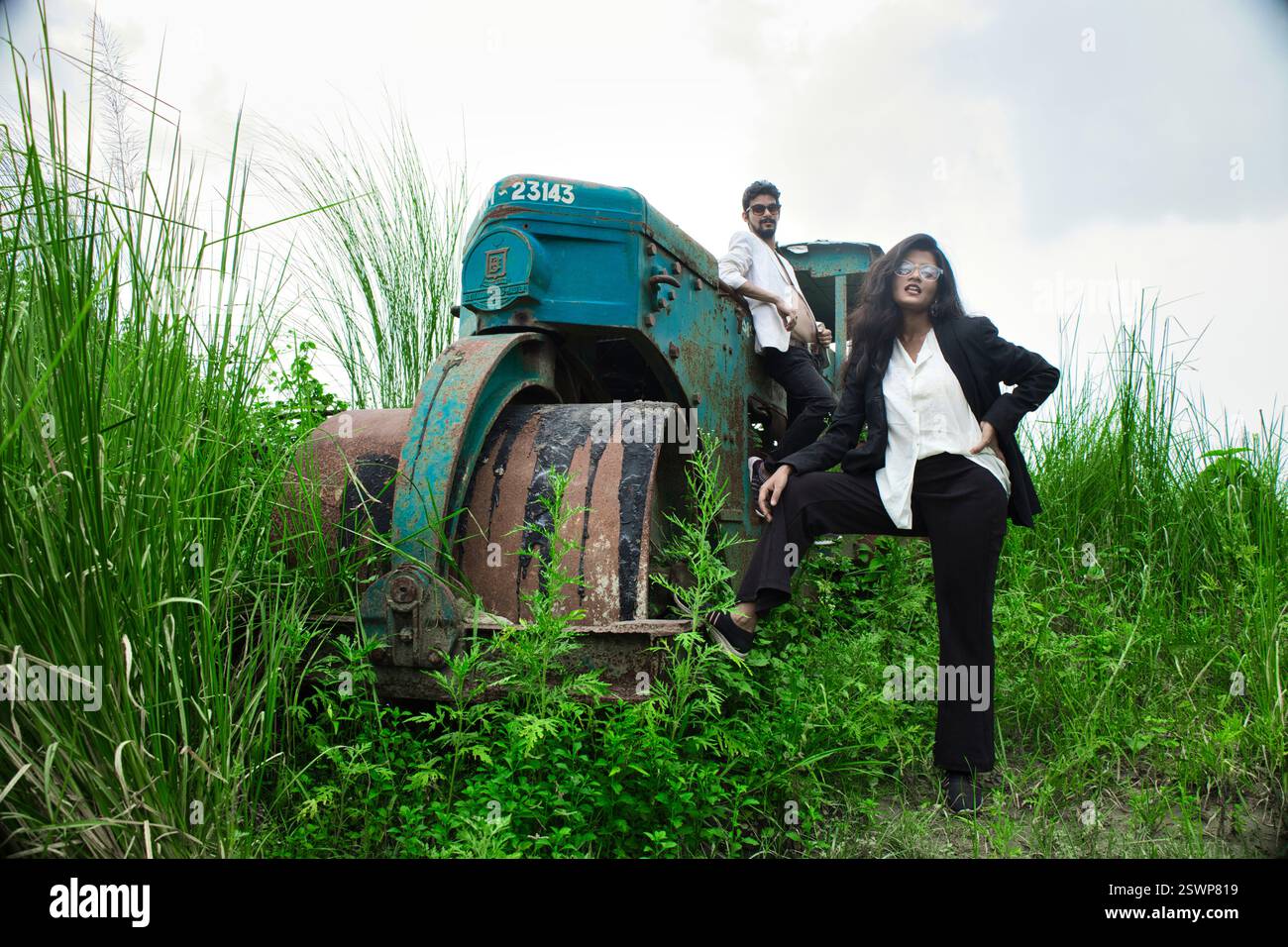 Modelli in posa in un campo aperto di fronte a un vecchio rullo. Il campo ha l'erba verde e sono vestiti in abiti bianchi e neri. Foto Stock