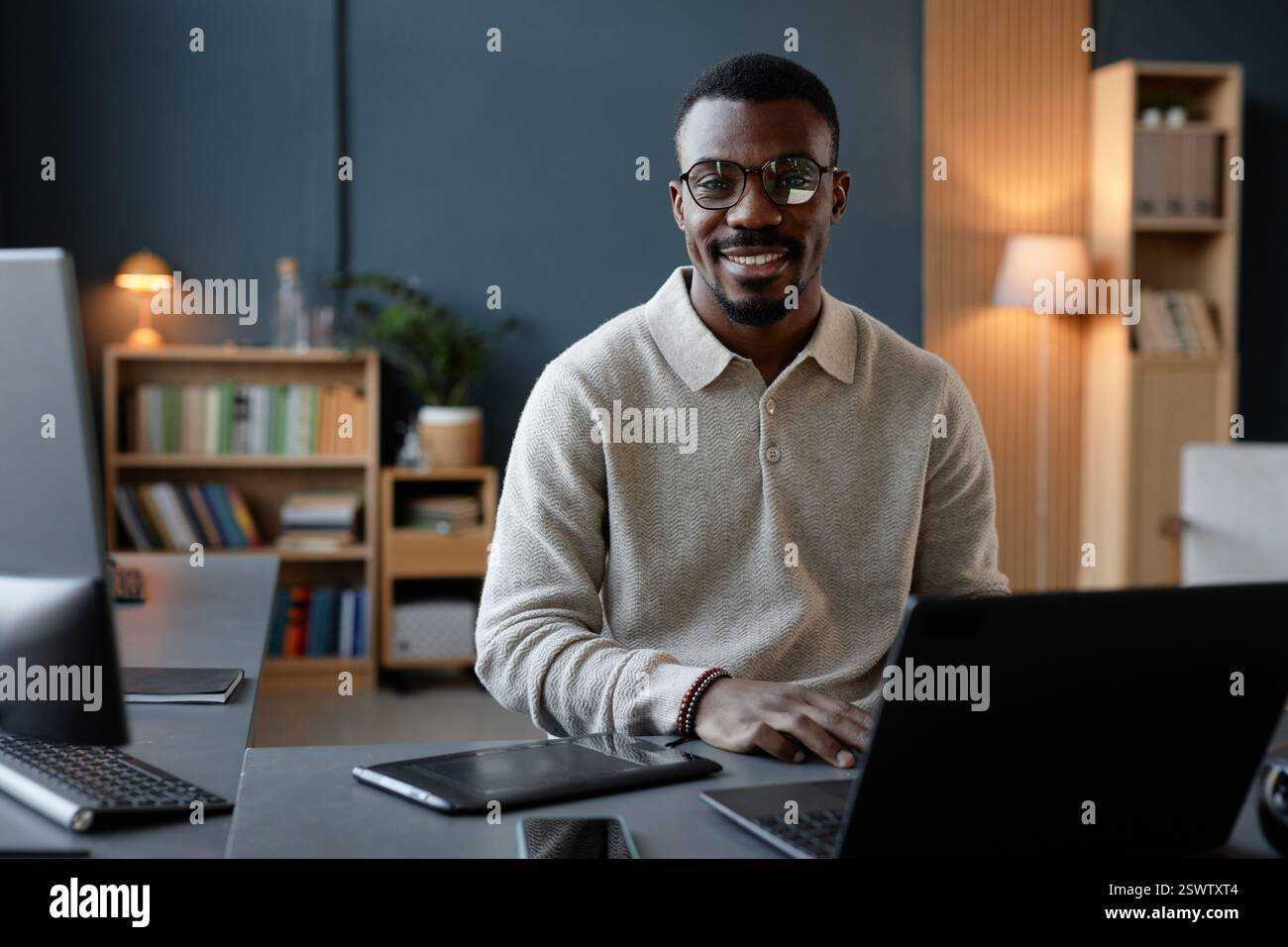 Ritratto di Cheerful Black, stilista maschile con occhiali che indossano una felpa da polo beige in posa alla scrivania, ingombrante di dispositivi per l'arte digitale in ufficio Foto Stock
