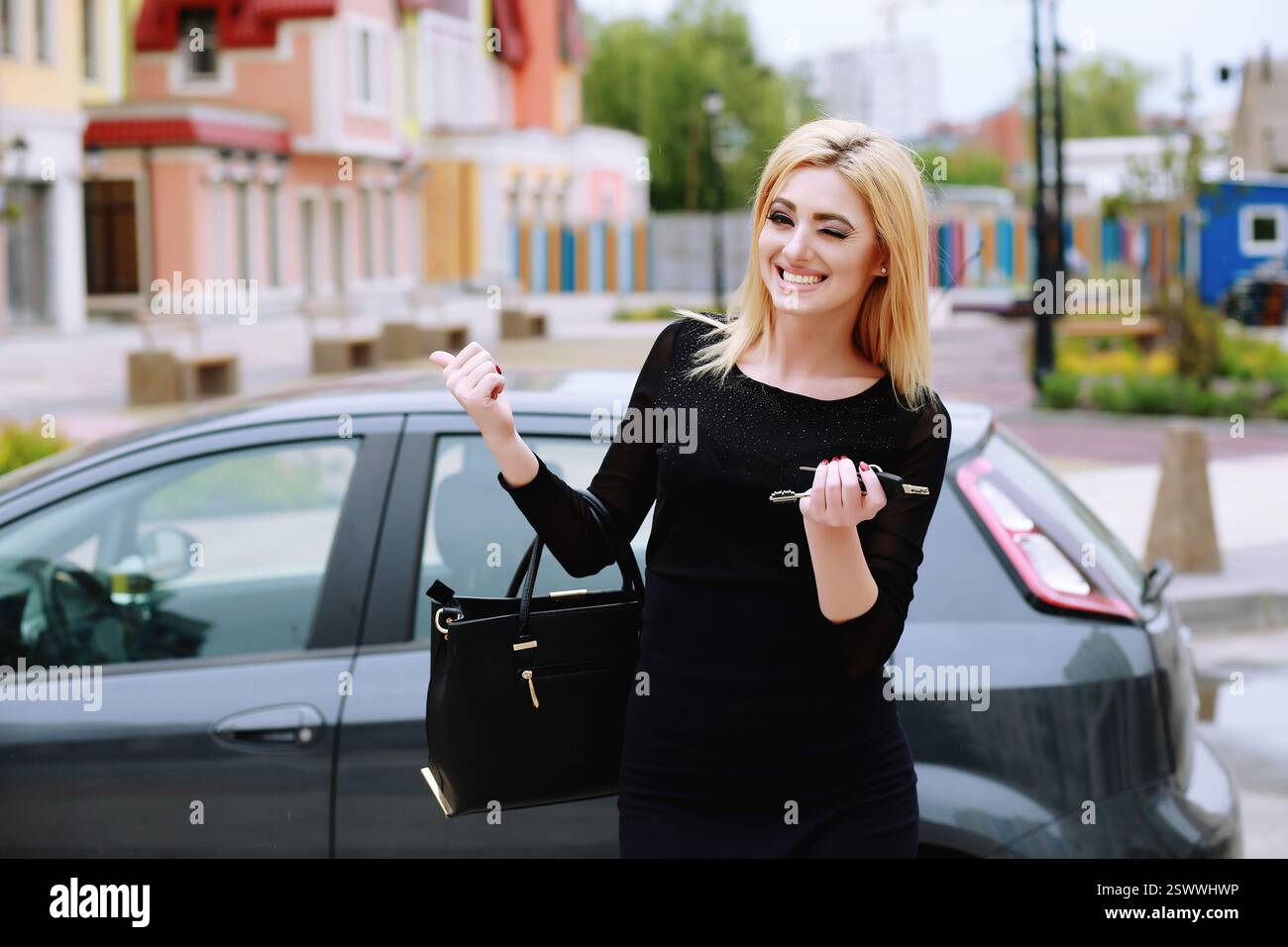 una bella donna sorride e sorride, indicando la macchina. Elegante signora in piedi vicino all'auto e con le chiavi dell'auto, all'aperto Foto Stock