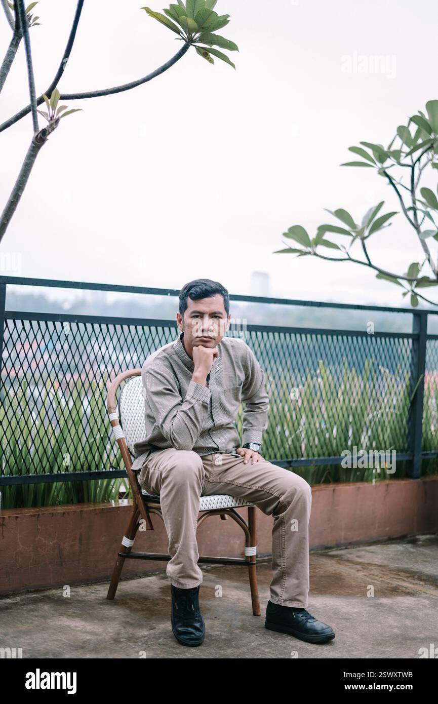 Un uomo premuroso è seduto su una sedia a contemplare mentre osserva il paesaggio urbano. Sembra penoso nella sua posizione, trovando un momento di pace in mezzo a te Foto Stock