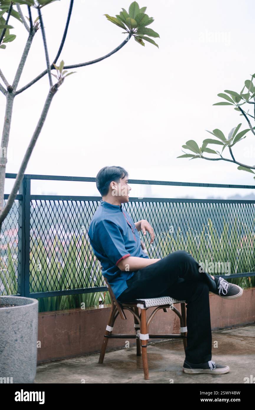 Un uomo asiatico siede su una sedia all'aperto, perso nella contemplazione e godendo dell'atmosfera tranquilla. La scena trasuda tranquillità e serenità, offrendo Foto Stock