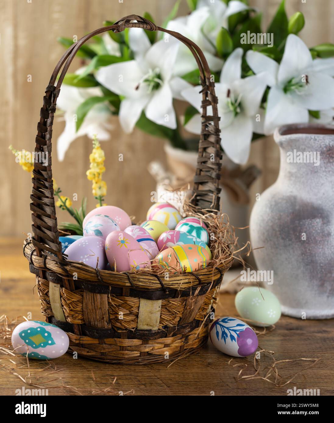 Uova di Pasqua decorate in un cestino con gigli bianchi su fondo rustico in legno Foto Stock