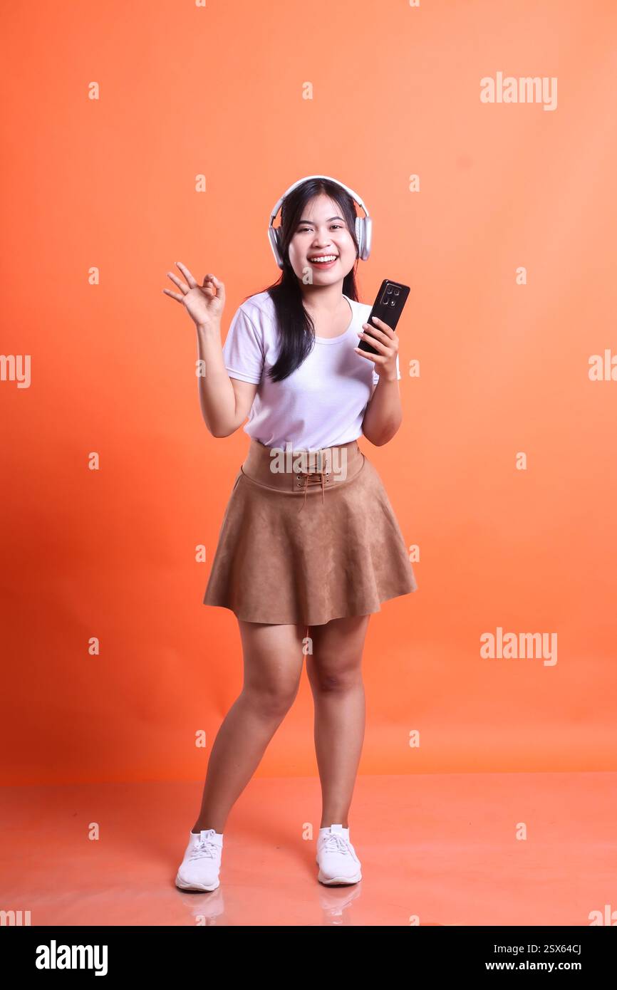 Una donna asiatica ascolta la musica con le cuffie e tiene in mano uno smartphone sorridente. Indossa una gonna e una camicia, isolata sul retro arancione Foto Stock