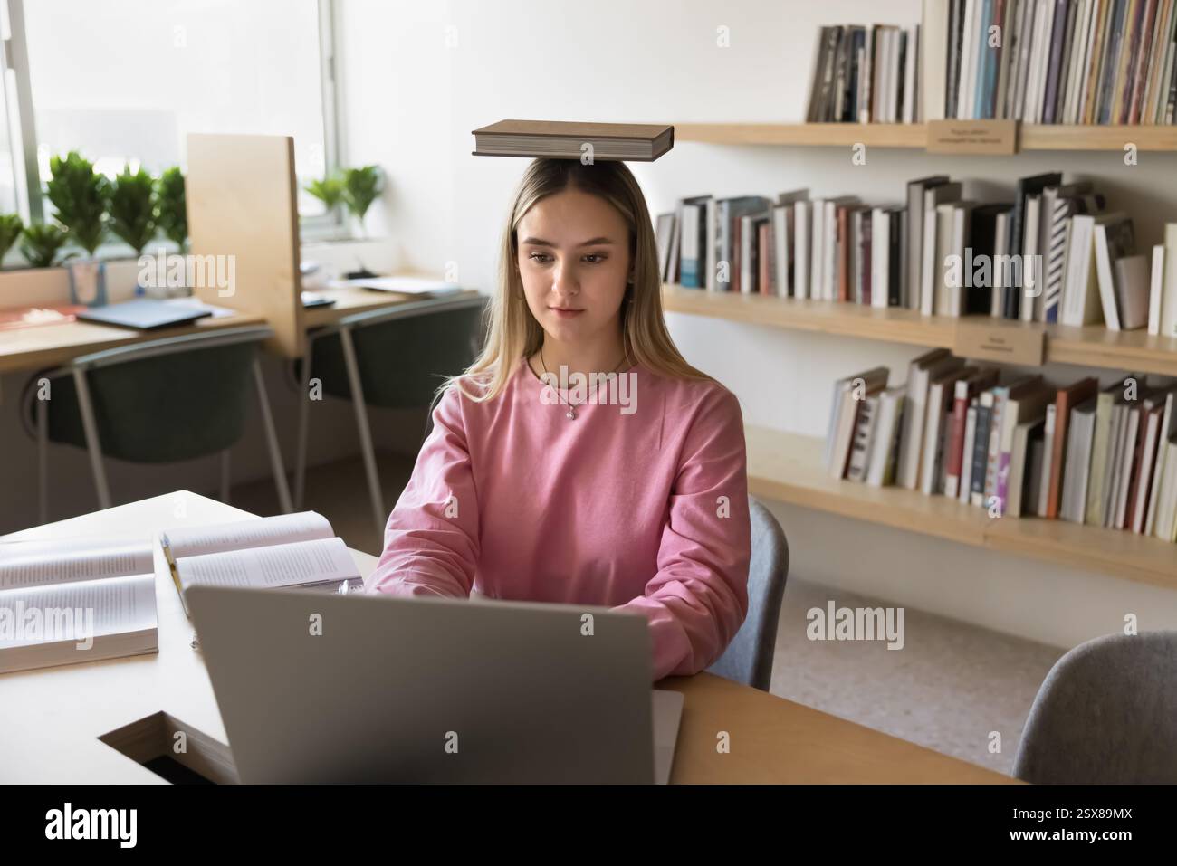 Ragazza che studia in biblioteca con un libro equilibrato sulla testa Foto Stock
