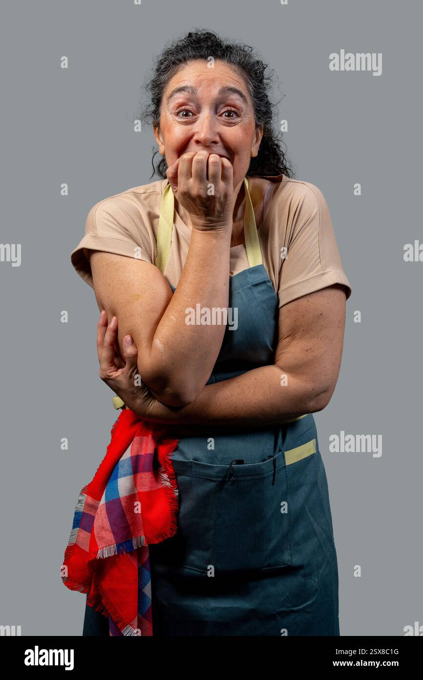 Chef donna anziana con grembiule e asciugamano da cucina rosso e blu che le mordono le unghie, mostrando ansia e nervosismo su sfondo grigio Foto Stock