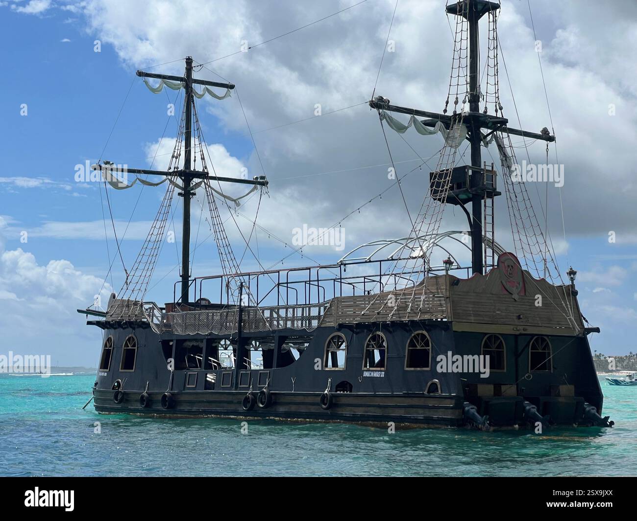 Una nave in legno a tema pirata con alberi alti e sartiame, che galleggia sulle acque turchesi sotto un cielo parzialmente nuvoloso, evocando avventura marittima e istore - Immagine stock catturata con smartphone