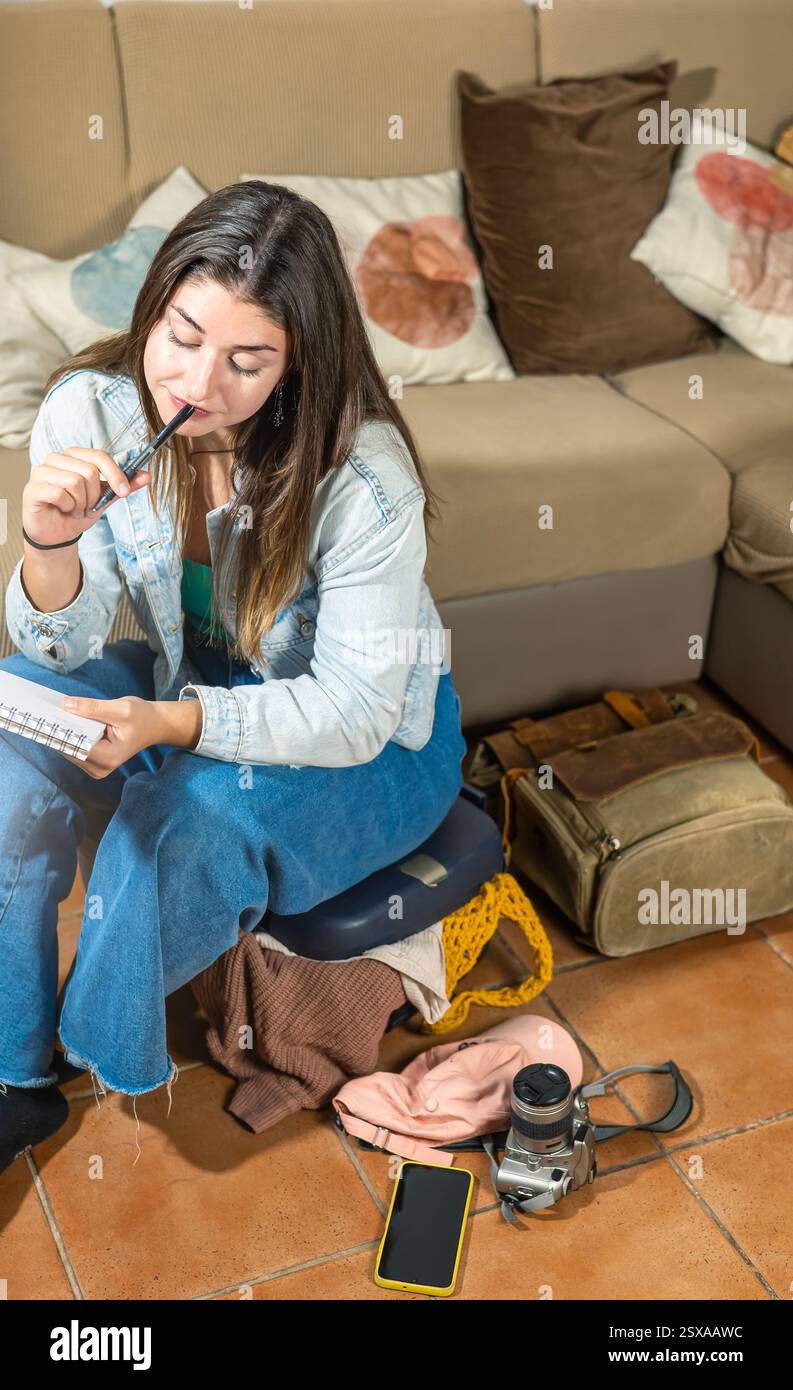 Giovane viaggiatrice seduto sulla sua valigia, concentrandosi sul suo notebook mentre esamina l'elenco delle cose che deve portare. Circa h Foto Stock