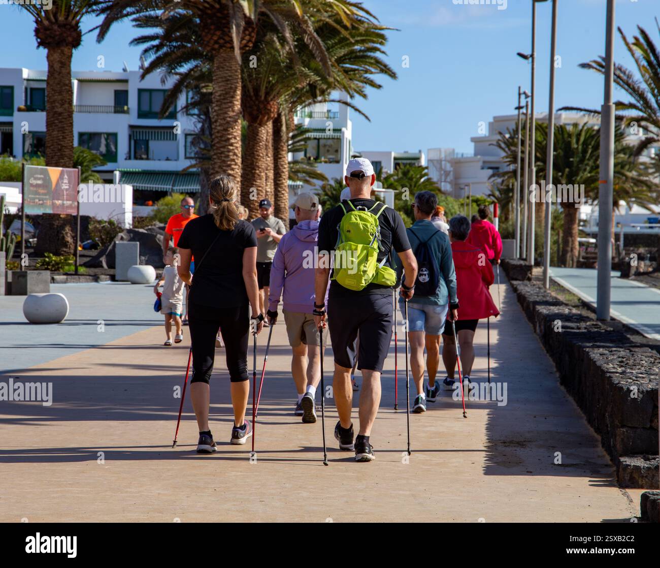 Gruppo di persone che camminano con i bastoncini nordici lungo una passeggiata fiancheggiata da palme in una cittadina costiera soleggiata Costa Teguise Lanzarote Foto Stock