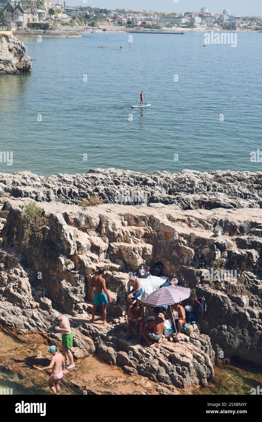 Un gruppo di amici si rilassa sulle rive rocciose della spiaggia di Santa Marta, mentre un paddleboarder scivola sullo sfondo Foto Stock