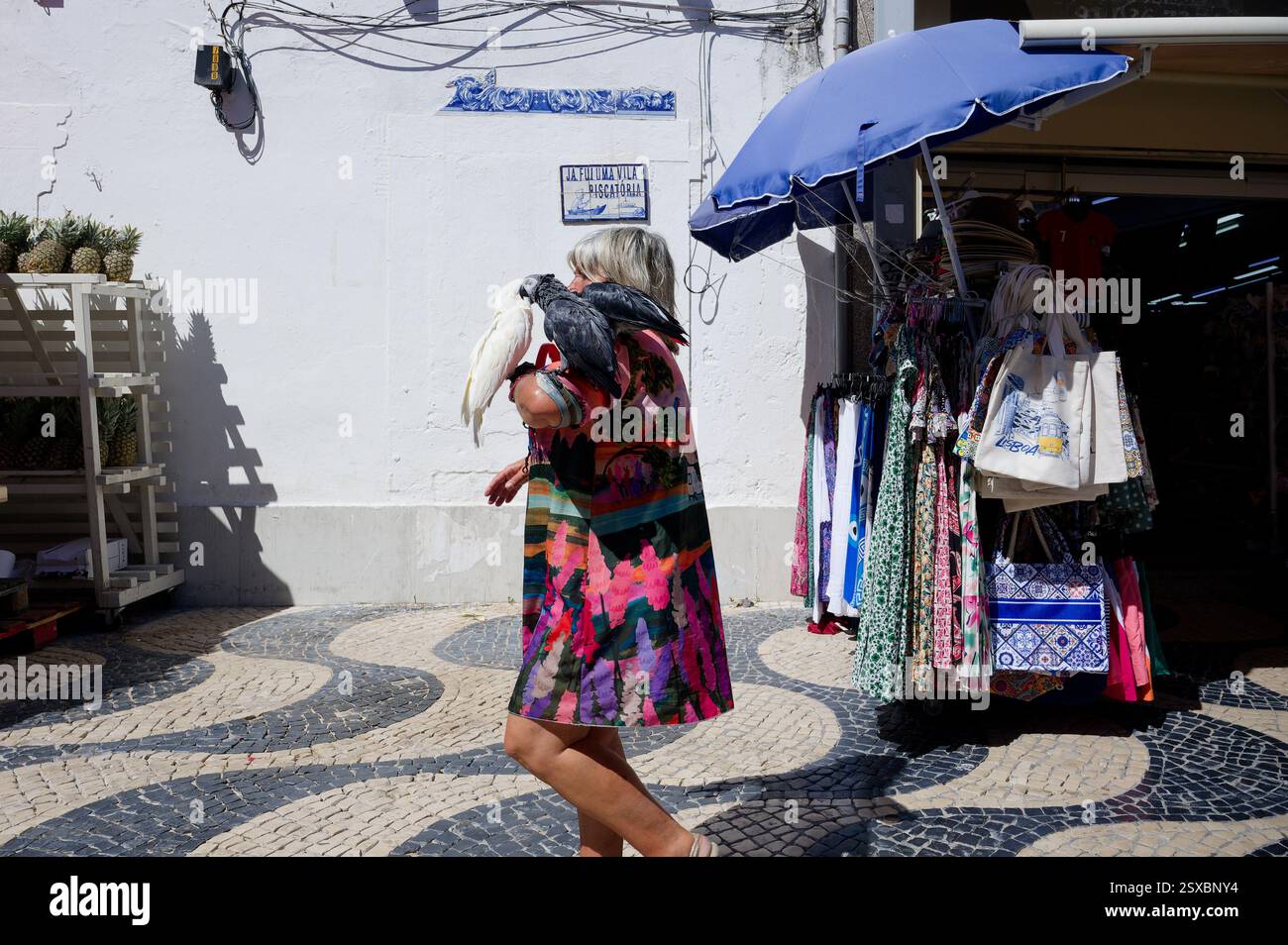 Una donna con un vestito colorato cammina per le strade di Cascais con due pappagalli sul braccio. Foto Stock Una donna con un vestito colorato cammina per le strade di Cascais con due pappagalli sul braccio. Foto Stock