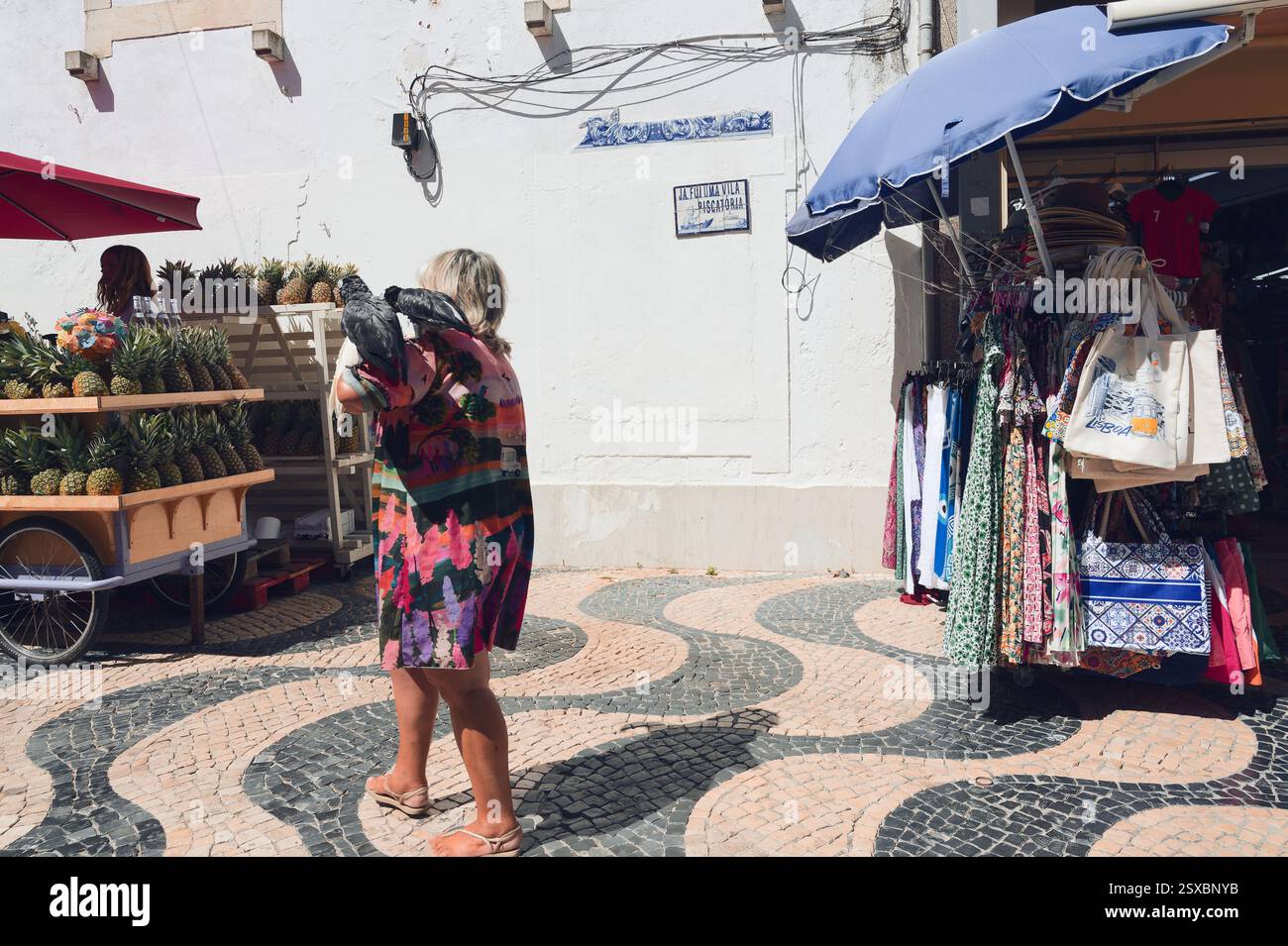 Una donna con un vestito colorato cammina per le strade di Cascais con due pappagalli sul braccio. Foto Stock