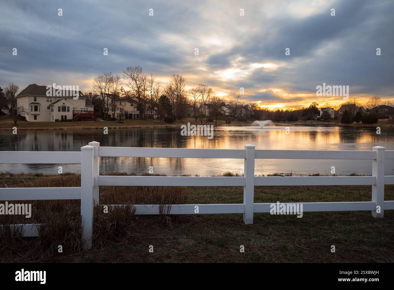 Uno splendido tramonto invernale proietta colori caldi su uno stagno ghiacciato con una fontana d'acqua, mentre una recinzione bianca in primo piano aggiunge fascino alla tranquillità Foto Stock