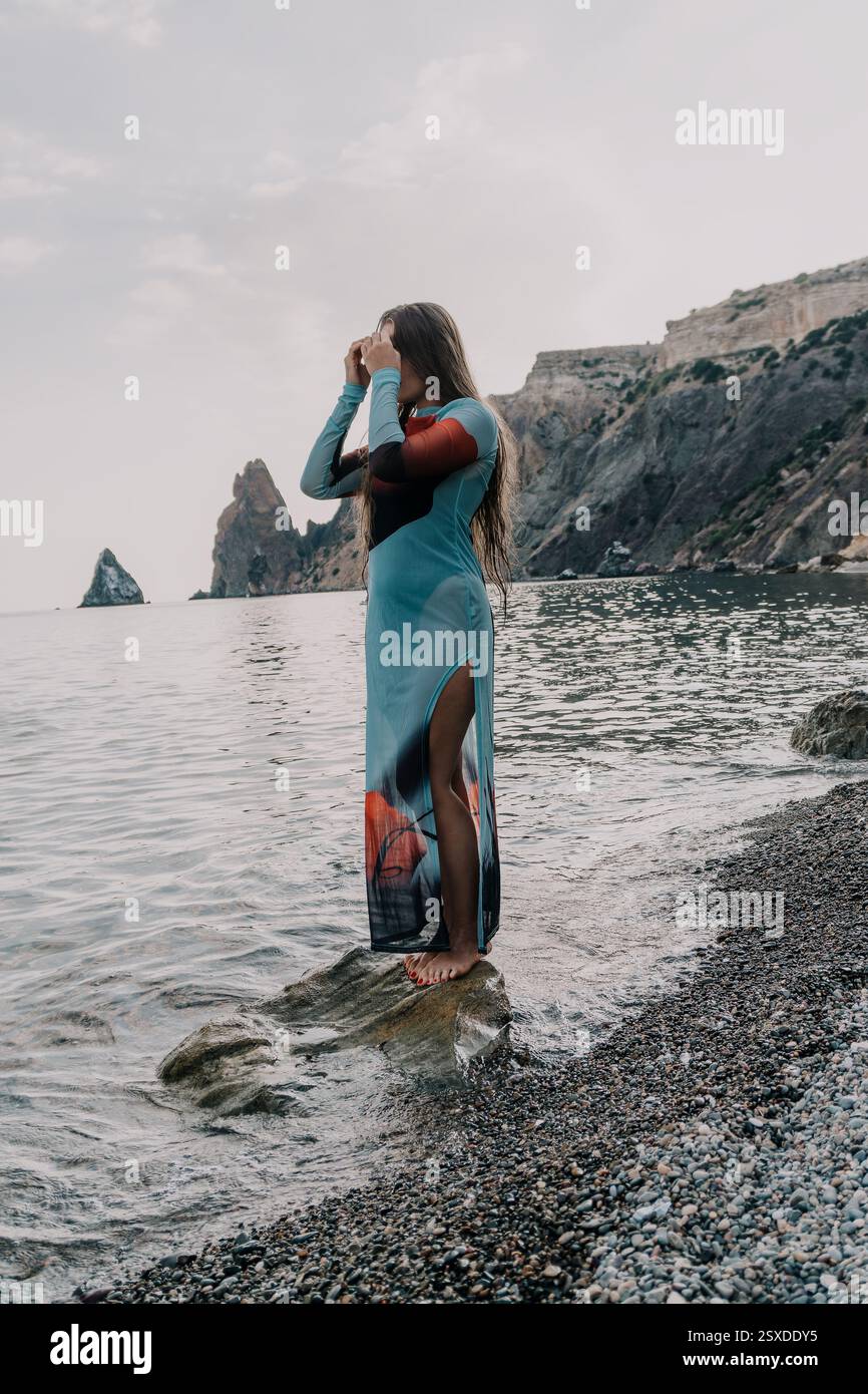 Woman Dress Beach Seascape - Una donna con un abito lungo si erge su una spiaggia rocciosa che si affaccia sul mare. Foto Stock